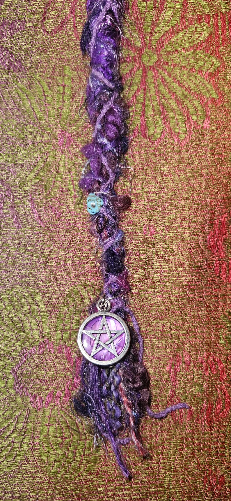 Puede incluir: Una envoltura de rastas morada hecha a mano con un dije de pentagrama plateado. La envoltura est&aacute; hecha de varios tonos de hilo morado y presenta una peque&ntilde;a cuenta turquesa. El fondo es una tela estampada en tonos verdes y rosas.