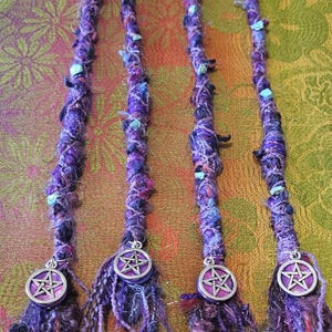 Puede incluir: Cuatro extensiones de rastas moradas con bandas el&aacute;sticas negras. Las rastas est&aacute;n adornadas con cuentas azules y verdes y amuletos de pent&aacute;culo plateados. Las extensiones est&aacute;n hechas de fibras moradas y negras.