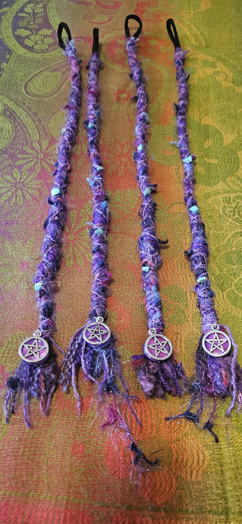 Puede incluir: Cuatro extensiones de rastas moradas con bandas el&aacute;sticas negras. Las rastas est&aacute;n adornadas con cuentas azules y verdes y amuletos de pent&aacute;culo plateados. Las extensiones est&aacute;n hechas de fibras moradas y negras.