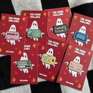May include: Set of ten enamel pins featuring book club themes. The pins are shaped like tickets and feature various book club names, such as "Spooky Book Club", "True Crime", and "Romance Book Club". The pins are on red cards with a ghost illustration and the text "The Spine Chillers".