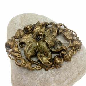 May include: A vintage brass brooch featuring a detailed floral design. The brooch showcases a large flower at the center, surrounded by leaves and buds, all in a warm, antique gold color. The pin is oval-shaped and likely made of metal.