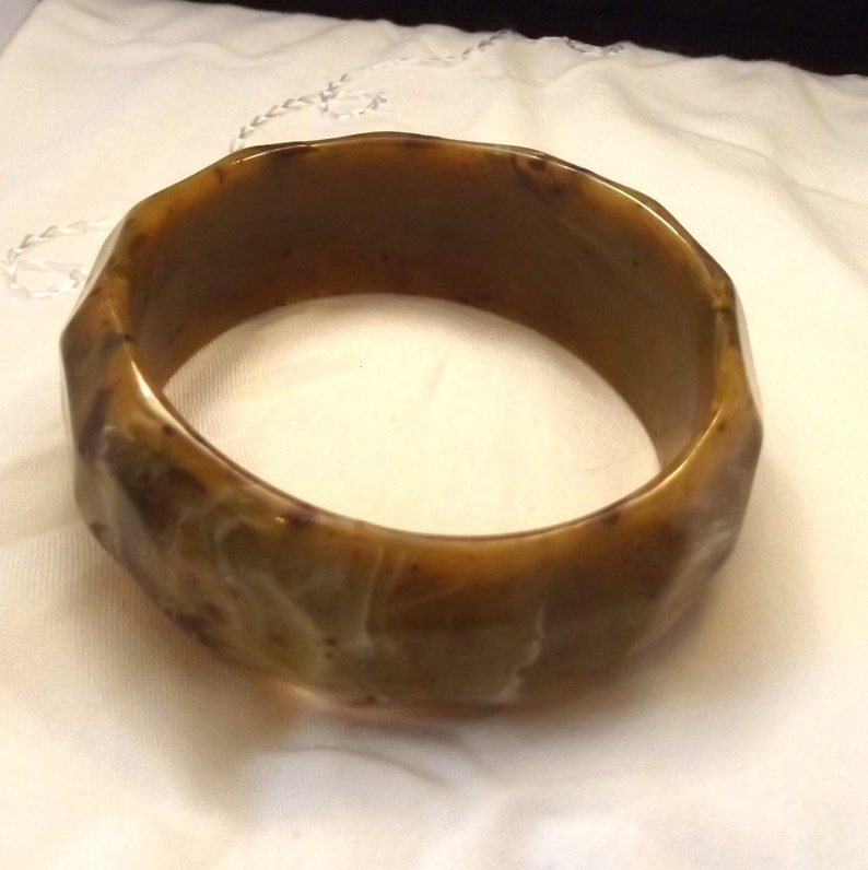 K&ouml;nnte beinhalten: Ein facettiertes, olivgr&uuml;nes Armreif-Armband. Das Armband hat ein geometrisches Design mit mehreren Seiten. Die Farbe ist eine Mischung aus Olivgr&uuml;n und Braun, wobei einige Bereiche durchscheinend erscheinen. Das Armband besteht aus einem festen Material.