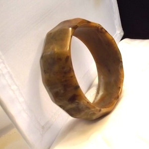 K&ouml;nnte beinhalten: Ein facettiertes, bernsteinfarbenes Armreif-Armband. Das Armband hat ein geometrisches Design mit mehreren flachen Seiten. Die Farbe ist eine Mischung aus hellem und dunklem Bernstein mit einigen dunkleren braunen Flecken. Das Armband besteht aus einem glatten, polierten Material.