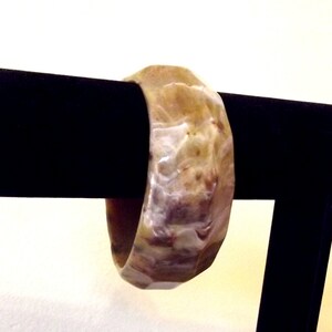 K&ouml;nnte beinhalten: Ein marmoriertes, beige-braunes Armreif-Armband. Das Armband ist dick und hat eine abgerundete, geometrische Form. Die Farben wirbeln zusammen und erzeugen ein nat&uuml;rliches, organisches Muster. Das Armband wird auf einem schwarzen St&auml;nder pr&auml;sentiert.