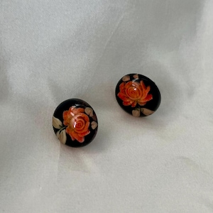 May include: Two black oval-shaped earrings. Each earring features a vibrant orange rose with green leaves and a cream-colored background. The earrings are set against a white background.