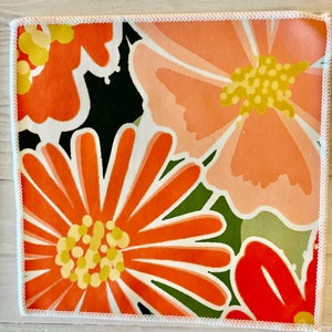 May include: A square fabric napkin with a white border and a floral pattern in shades of orange, pink, and green.