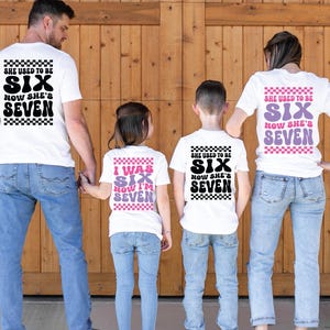 May include: A family of four wearing white t-shirts with the text "SHE USED TO BE SIX NOW SHE'S SEVEN" in black and purple. The shirts have a checkered pattern. The family is standing in front of a wooden door.