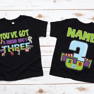 以下が含まれることがあります： カラフルなグラフィックが施された2枚の黒いTシャツ。1枚のシャツには、漫画のキャラクターと一緒に「You've got a friend who's THREE」と書かれています。もう1枚のシャツには、大きな数字の「3」と緑色の「NAME」という文字が書かれています。
