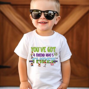 以下が含まれることがあります： サングラスをかけた子供が、"YOU'VE GOT A FRIEND WHO'S THREE"と書かれた白いTシャツ、茶色のショートパンツ、白いスニーカーを着用しています。 子供は笑顔で、木のドアの前に立っています。