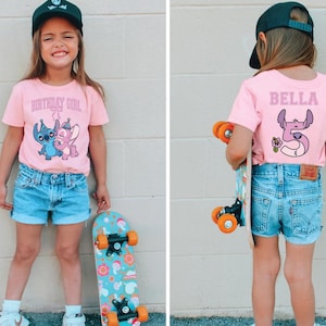 May include: A young girl wearing a pink t-shirt with the text "BIRTHDAY GIRL" and a cartoon graphic of Stitch and Angel. She is also wearing denim shorts, a black cap, and holding a skateboard with a colorful design.