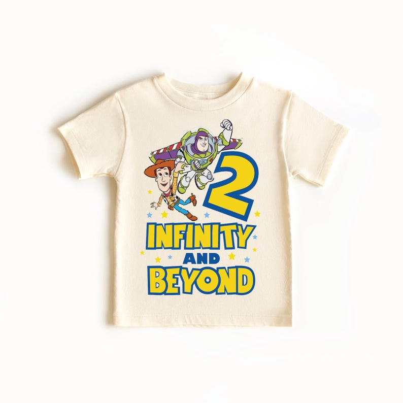 Op de afbeelding: Een cr&egrave;mekleurig T-shirt met een cartoon afbeelding van Woody en Buzz Lightyear uit Toy Story. Het shirt heeft een blauw nummer "2" met gele omtrek en de tekst "Oneindig en verder" in blauw met gele omtrek.