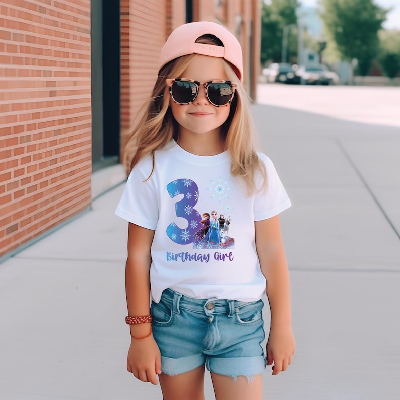 Pode incluir: Uma camiseta branca com um desenho azul do n&uacute;mero 3, flocos de neve e personagens do filme Frozen. O texto "Birthday Girl" est&aacute; abaixo. A menina est&aacute; usando shorts jeans, &oacute;culos de sol e um bon&eacute; rosa.