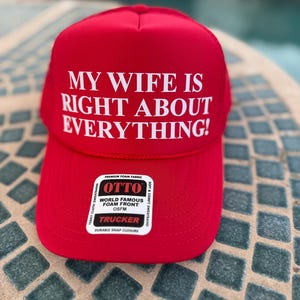 May include: A red trucker hat with the text "MY WIFE IS RIGHT ABOUT EVERYTHING!" in white. The hat has a foam front and a snap closure. The brand label is visible.