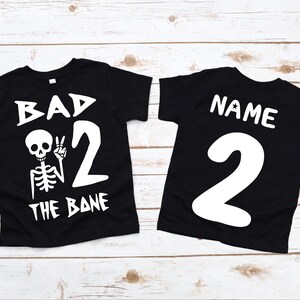 Pode incluir: Duas camisetas pretas com texto e gráficos brancos. A primeira camiseta diz "Bad to the Bone" com um esqueleto levantando dois dedos. A segunda camiseta diz "Name" e tem o número "2" nela.