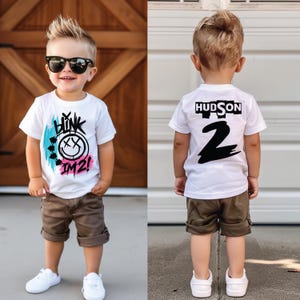 May include: A white short-sleeve t-shirt featuring a graphic print with the text "BLINK IM 2!" in black, accented by blue and pink brushstrokes. The back of the shirt displays the name "HUDSON" above a large black number "2" in a bold font.