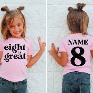 May include: Pink t-shirt with the text "eight is great" in black. The back of the shirt has the word "NAME" and the number "8" in black. The shirt is worn by a child.