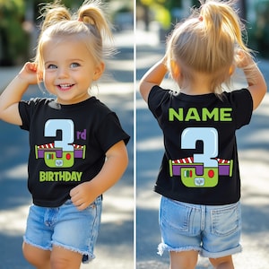 Op de afbeelding: Een zwart T-shirt met een groene, paarse en rode afbeelding van het getal 2 met de tekst "3rd Birthday" eronder. De achterkant van het shirt heeft het getal 2 en het woord "NAME" erboven.