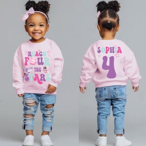 Puede incluir: Una sudadera rosa con el texto "REACH FOUR THE STARS" y un diseño espacial. La parte trasera de la sudadera tiene el nombre "SOPHIA" y el número "4". Combinada con vaqueros azules rotos.