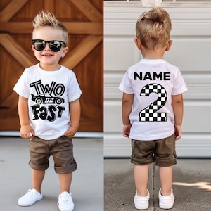 May include: A white short-sleeved t-shirt featuring a black graphic print. The front displays the text "TWO FAST" with a race car illustration. The back features the text "NAME" above a large number "2" with a black and white checkered pattern.