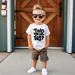 May include: A white short-sleeved t-shirt featuring the black text "TWO FAST" and a graphic of a race car with the number 02. The shirt is paired with brown shorts and white sneakers. The wearer is also sporting black sunglasses.