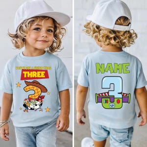 May include: Light blue t-shirt with a graphic featuring characters from Toy Story. The front of the shirt says "YOU'VE GOT A FRIEND WHOS THREE" with a large number 3. The back has "NAME" above a number 3 with a Buzz Lightyear design.