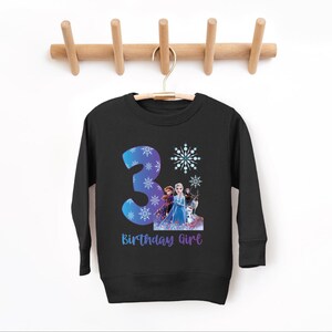 Pode incluir: Camisola preta de manga comprida com um n&uacute;mero 3 azul e roxo e detalhes de flocos de neve. A camisola tem um gr&aacute;fico de personagens animados e o texto "Birthday Girl" em roxo.