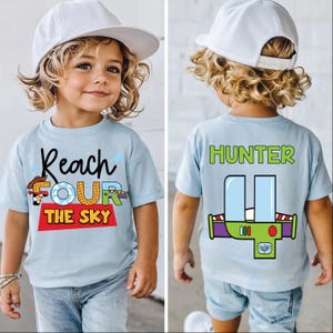 May include: A pale blue t-shirt featuring the text "Reach FOUR The Sky" and a cartoon graphic. The back of the shirt displays the name "HUNTER" and a spaceship graphic. The t-shirt is worn by a young child.