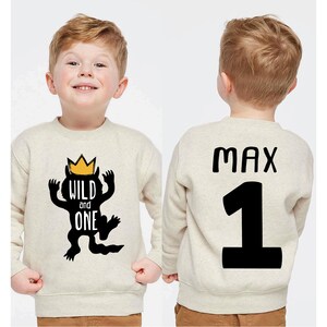 May include: A beige sweatshirt with a black graphic of a monster wearing a gold crown and the words "WILD and ONE". The back of the sweatshirt has the name "MAX" and the number "1" in black. The sweatshirt is for a child.