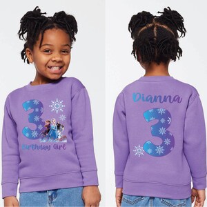 Pode incluir: Um moletom lil&aacute;s com um grande n&uacute;mero 3 e desenhos de flocos de neve, al&eacute;m de um gr&aacute;fico de Frozen. A frente diz "Birthday Girl". A parte de tr&aacute;s do moletom tem o nome "Dianna" e o n&uacute;mero 3.