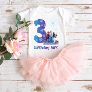 Pode incluir: Um body branco para beb&eacute; com um grande n&uacute;mero 3 azul, flocos de neve e um gr&aacute;fico de personagens. O texto "Birthday Girl" est&aacute; abaixo do n&uacute;mero. Uma saia de tule rosa &eacute; combinada com o body. O conjunto est&aacute; numa superf&iacute;cie de madeira branca.