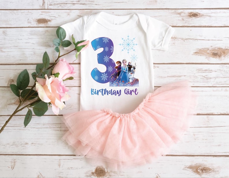 Pode incluir: Um body branco para beb&eacute; com um grande n&uacute;mero 3 azul, flocos de neve e um gr&aacute;fico de personagens. O texto "Birthday Girl" est&aacute; abaixo do n&uacute;mero. Uma saia de tule rosa &eacute; combinada com o body. O conjunto est&aacute; numa superf&iacute;cie de madeira branca.