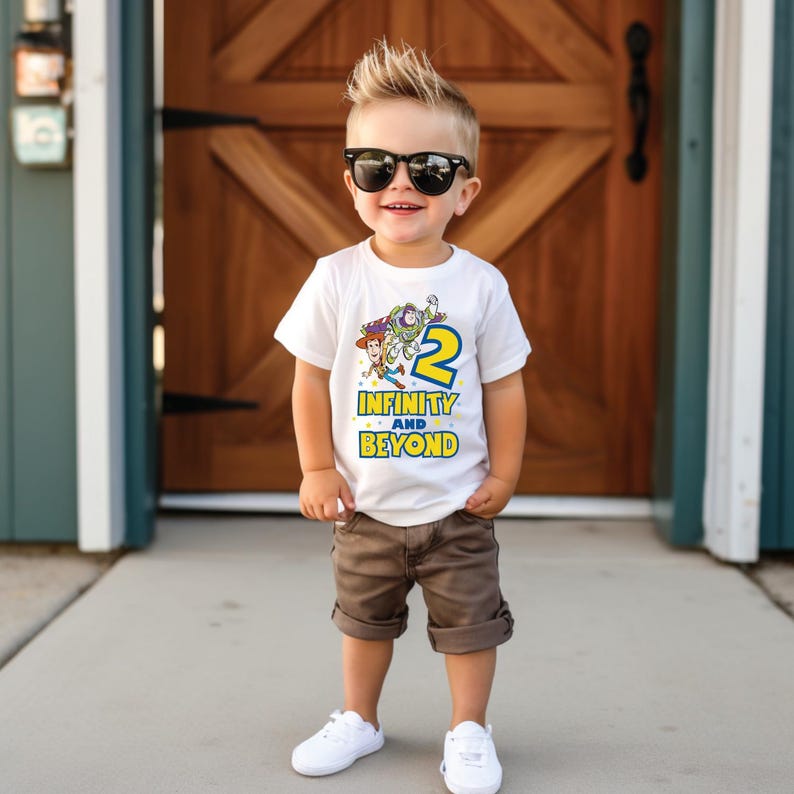 Op de afbeelding: Een wit T-shirt met een Toy Story-thema met Woody, Buzz Lightyear en het nummer 2. Op het shirt staat de tekst "Infinity and Beyond". Een jonge jongen draagt het shirt en glimlacht.
