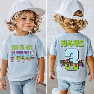 以下が含まれることがあります： 「YOU'VE GOT A FRIEND WHO'S THREE」の文字と漫画のキャラクターがプリントされた水色のTシャツ。背中には「NAME」の文字と、バズ・ライトイヤーのデザインの大きな数字「3」がプリントされています。子供がTシャツを着ています。