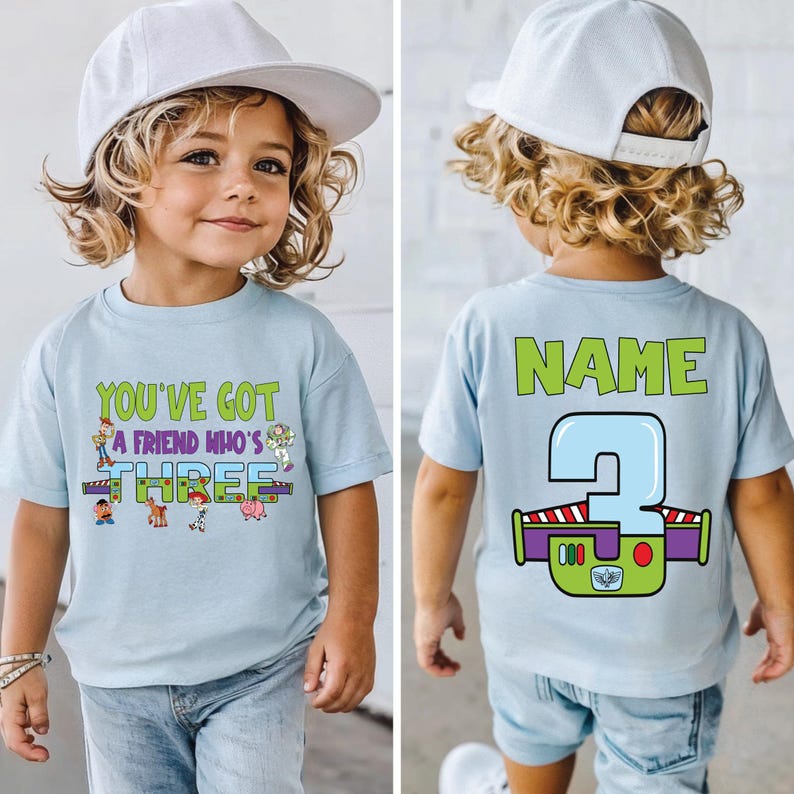 以下が含まれることがあります： 「YOU'VE GOT A FRIEND WHO'S THREE」の文字と漫画のキャラクターがプリントされた水色のTシャツ。背中には「NAME」の文字と、バズ・ライトイヤーのデザインの大きな数字「3」がプリントされています。子供がTシャツを着ています。