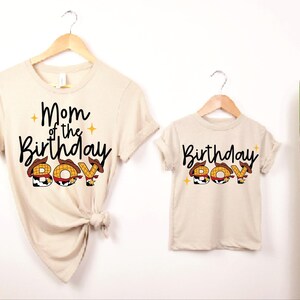 May include: Two beige t-shirts, one adult and one child size, hanging on wooden hangers. The adult shirt reads "Mom of the Birthday Boy" and the child's shirt says "Birthday Boy". The word "BOY" is designed with a cowboy hat theme.