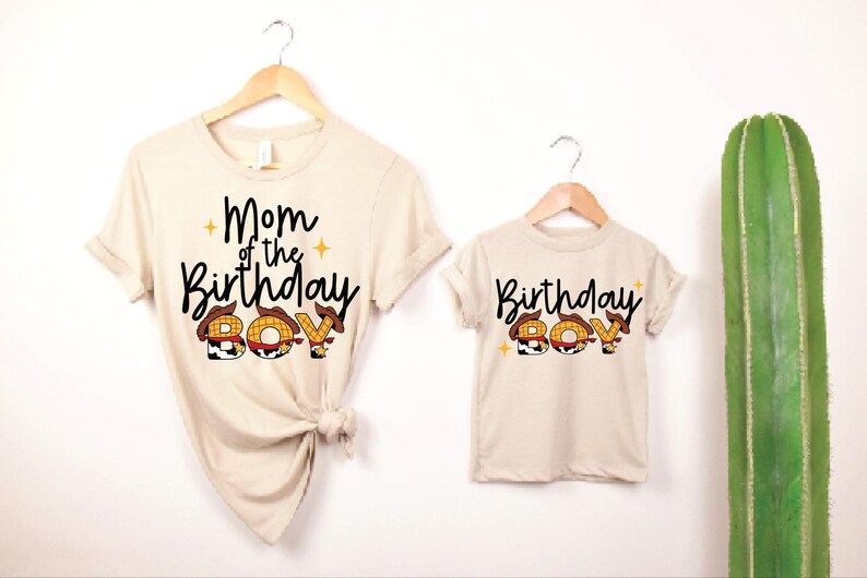 May include: Two beige t-shirts, one adult and one child size, hanging on wooden hangers. The adult shirt reads "Mom of the Birthday Boy" and the child's shirt says "Birthday Boy". The word "BOY" is designed with a cowboy hat theme.