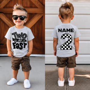May include: A light gray t-shirt featuring the text TWO FAST with a race car graphic on the front and the word NAME above a large checkered number 2 on the back. The shirt is paired with brown shorts and white sneakers for a complete outfit.