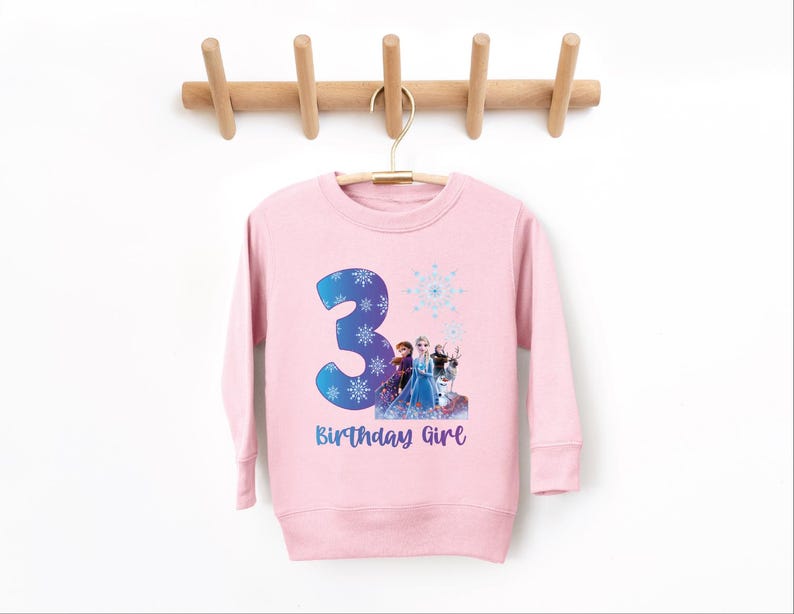 Pode incluir: Camisola rosa claro para crian&ccedil;a com o n&uacute;mero 3 azul, flocos de neve e uma imagem de personagens. O texto "Birthday Girl" est&aacute; em baixo. Pendurada num cabide de madeira.