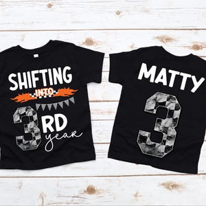 May include: Two black t-shirts with white text and a checkered number 3. One shirt says "Shifting into 3rd year" with orange flames and a checkered flag design. The other shirt says "Matty" above the number 3.