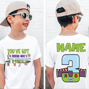 以下が含まれることがあります： 「YOU'VE GOT A FRIEND WHO'S THREE」の文字とトイ・ストーリーのキャラクターがプリントされた白いTシャツ。Tシャツの背面には「NAME」の文字と、バズ・ライトイヤーのデザインの大きな数字3が描かれています。男の子は帽子とサングラスを着用しています。