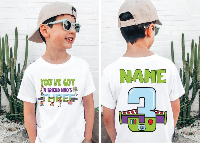 以下が含まれることがあります： 「YOU'VE GOT A FRIEND WHO'S THREE」の文字とトイ・ストーリーのキャラクターがプリントされた白いTシャツ。Tシャツの背面には「NAME」の文字と、バズ・ライトイヤーのデザインの大きな数字3が描かれています。男の子は帽子とサングラスを着用しています。
