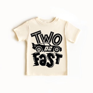 May include: A cream-colored short-sleeve t-shirt featuring a black graphic print. The design includes the text "TWO" above a race car illustration with the number "02" on its side, followed by the word "FAST" in a bold, stylized font below.