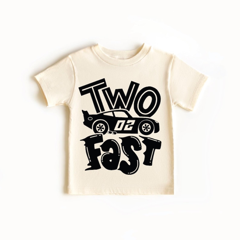 May include: A cream-colored short-sleeve t-shirt featuring a black graphic print. The design includes the text "TWO" above a race car illustration with the number "02" on its side, followed by the word "FAST" in a bold, stylized font below.