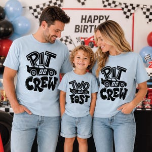 May include: Light blue t-shirts featuring black graphic prints. Two shirts display the text PIT CREW with a race car illustration labeled 02. One shirt displays the text TWO FAST with a race car illustration labeled 02. The shirts are short-sleeved.