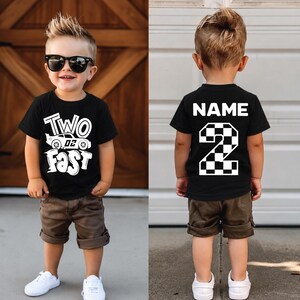 May include: A black short-sleeved t-shirt featuring the white text "TWO FAST" with a race car graphic on the front and the white text "NAME" above a large checkered number "2" on the back, paired with brown shorts and white sneakers.