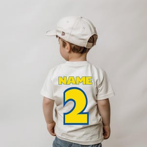Op de afbeelding: Een wit T-shirt met een geel nummer "2" en het woord "NAME" in gele letters op de achterkant. Het shirt wordt gedragen door een kind dat een witte baseballpet draagt.