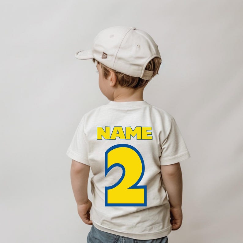Op de afbeelding: Een wit T-shirt met een geel nummer "2" en het woord "NAME" in gele letters op de achterkant. Het shirt wordt gedragen door een kind dat een witte baseballpet draagt.