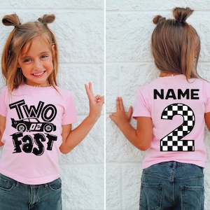 May include: A light pink short-sleeve t-shirt featuring a black graphic on the front with a race car and the text "TWO FAST". The back of the shirt displays the word "NAME" above a large number "2" with a black and white checkered pattern.