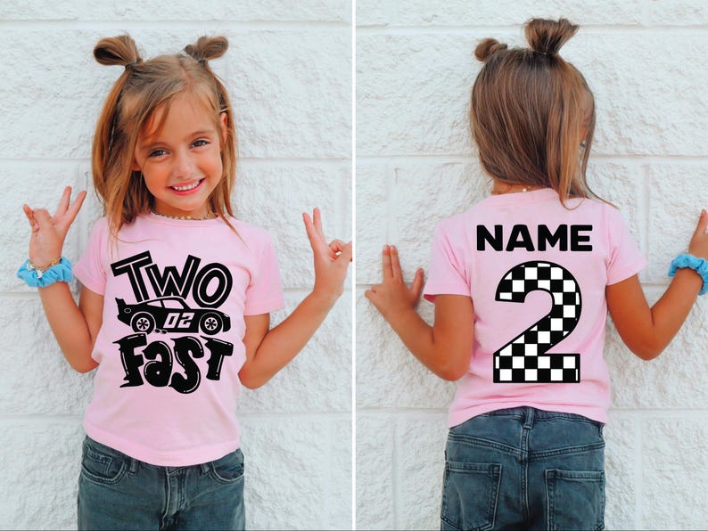 May include: A light pink short-sleeve t-shirt featuring a black graphic on the front with a race car and the text "TWO FAST". The back of the shirt displays the word "NAME" above a large number "2" with a black and white checkered pattern.