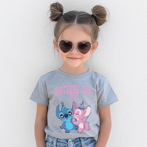 May include: A young girl wearing heart-shaped sunglasses and a gray t-shirt with the text "BIRTHDAY GIRL" in pink. The shirt features a graphic of two cartoon characters, one blue and one pink, wearing party hats.